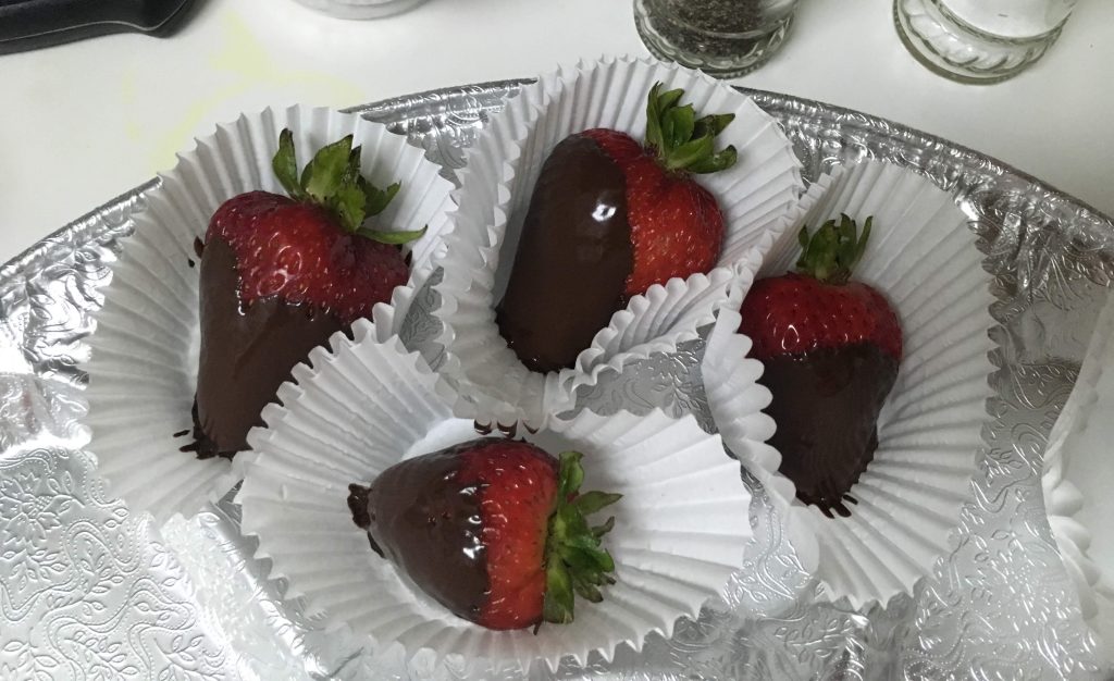 4 chocolate dipped strawberries in small paper cups set together on a glass plate.